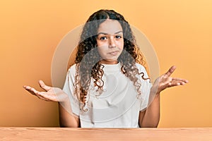 Teenager hispanic girl wearing casual clothes sitting on the table clueless and confused expression with arms and hands raised
