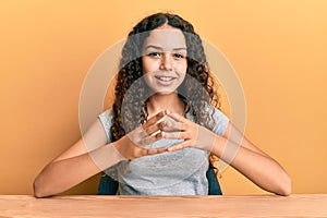 Teenager hispanic girl wearing casual clothes sitting on the table hands together and fingers crossed smiling relaxed and cheerful