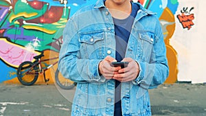 Teenager in a jeans jacket is standing with his mobile phone near a graffiti wall