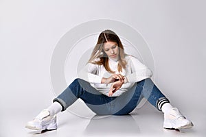 Teenager in jeans, sweater, socks and sneakers. She looking at her watch sitting on floor, legs apart,  on white. Close up