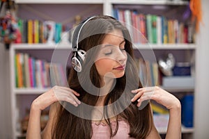 A teenager listens to music with her eyes closed and a bookcase with books in the tel.