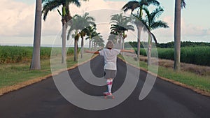 A teenager with long hair rides a skateboard along a beautiful road with palm trees. Los Angeles. Boy longboarder riding