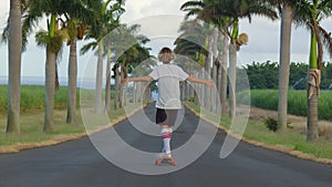 A teenager with long hair rides a skateboard along a beautiful road with palm trees. Los Angeles. Boy longboarder riding