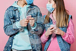 Teenager in medical mask holding smartphone near friend on pink background.