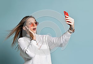 Teenager in orange sunglasses, watch, bracelet and sweater. She taking selfie, surprised, posing on blue background. Close up
