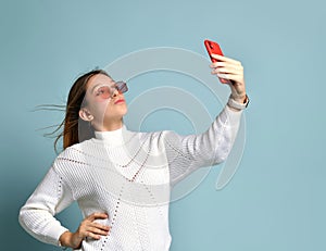 Teenager in orange sunglasses, watch, bracelet and sweater. She taking selfie, surprised, posing on blue background. Close up