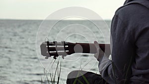 A teenager plays a guitar while sitting on the shore of a lake.