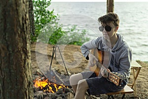 A teenager plays a guitar while sitting on the shore of a lake.