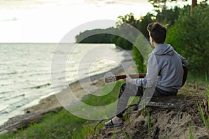 A teenager plays a guitar while sitting on the shore of a lake.