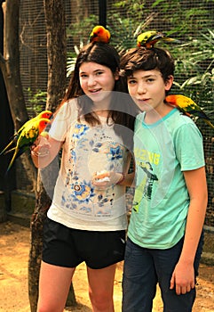 Teenager siblings boy and girl in open air zoo with parrots sitting on hand and head