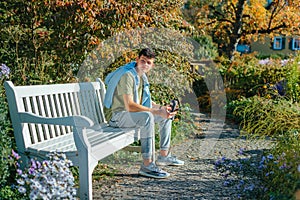 A Teenager Sits On A Bench In The Autumn Park Drinks Coffee From A Thermo Mug And Looks Into A Phone. Portrait Of