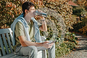 A Teenager Sits On A Bench In The Autumn Park Drinks Coffee From A Thermo Mug And Looks Into A Phone. Portrait Of