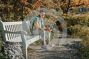 A Teenager Sits On A Bench In The Autumn Park Drinks Coffee From A Thermo Mug And Looks Into A Phone. Portrait Of