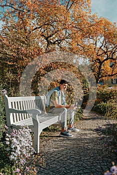 A Teenager Sits On A Bench In The Autumn Park Drinks Coffee From A Thermo Mug And Looks Into A Phone. Portrait Of