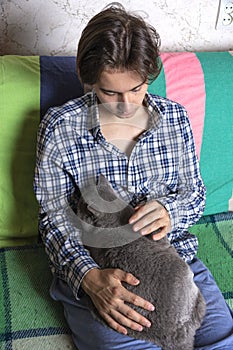 A teenager is sitting on the couch and stroking a Scottish cat sleeping on his lap