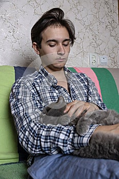 A teenager is sitting on the couch and stroking a Scottish cat sleeping on his lap