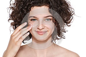 Teenager skincare. Beautiful teenage girl with gorgeous curly hair applying moisturiser face cream, looking at camera smiling.