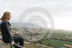 Teenager thinking sitting in fence on a viewpoint of Tenerife north. Blond hair girl relaxed with contemplation in distant in a