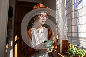 Teenager in vintage clothes standing in sunny room with cup of tea looking at camera with sly glance