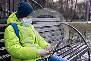 A teenager wearing a medical mask on a Park bench uses a smartphone and headphones.