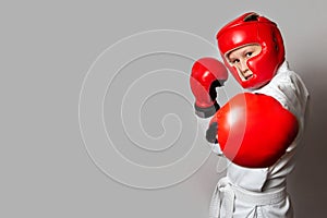 A teenager in a white kimono and gloves and a helmet for hand-to-hand combat on a gray background. Copy space. Sports exercises.