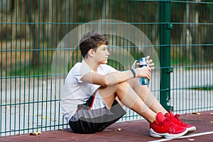 Teenager in a white t-shirt drinks water from bottle. Cute young boy on the street basketball playground in summer.