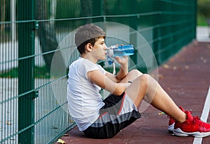 Teenager in a white t-shirt drinks water from bottle. Cute young boy on the street basketball playground in summer.