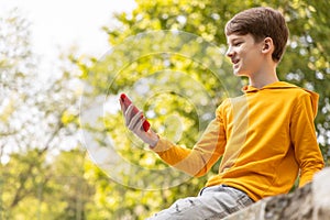 A teenager in a yellow hoodie, sitting in a park, smiles while looking at the screen of their smartphone.