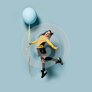 Teenager in yellow sweatshirt, black skirt, knee-highs, boots. She smiling, holding balloon, jumping up against blue background.