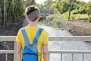A teenager in a yellow t-shirt with a blue backpack stands near the fence and looks at the river, the view from the back.