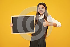 Teenager younf school girl holding school empty blackboard isolated on yellow background. Portrait of a teen female