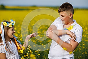 Teenagers: brother and sister with Ukrainian wreath with on head, in rapeseed field under blue sky