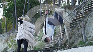 Teenagers in casual clothes dancing on the sandy bank of the river near the wooden stairs. Asian guy and caucasian girl