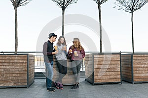 Teenagers eat street food, friends boy and two girls on city street with burgers and orange juice. City background, golden hour