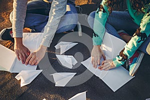 Teenagers making paper airplanes sitting at the roof of a building at sunset. Young people holding paper aeroplanes, love,