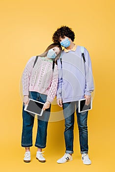 Teenagers in medical masks holding digital tablets on yellow background.