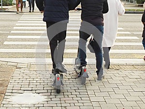 Teenagers on scooters cross the road along a pedestrian crossing among other people