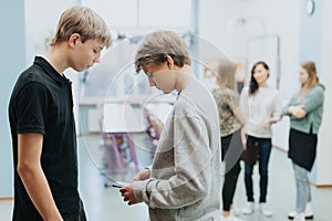 Teenagers are standing together at the side of the classroom, one of them is checking something on a mobile phone