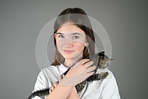 Teengirl holding a baby cat on gray background, cute tenderly smiling. Kitten sits on the female shoulder