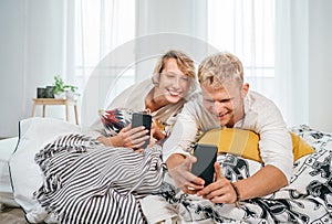 Teens couple lying in a cozy bed in the bedroom and browsing together internet or checking mails using a modern smartphones.