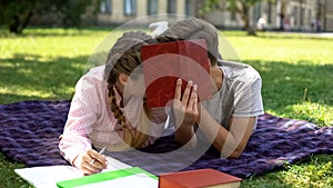 Teens hiding behind book and kissing, lying on plaid in park, first relations