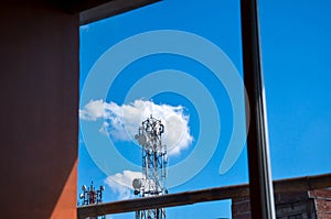 Telecommunication antenna or cell towers through a window against blue sky with floating clouds