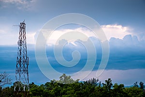 Telecommunication towers and green tree forest against blue cloudy sky with rays of light.