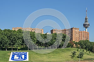Telecommunications tower of Madrid and in the foreground the trees of the park of Rome with the shield of the city in the grass