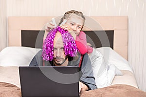 Telecommuting in quarantine. A fun family event. A little girl prevents dad from working at a computer. A child puts a bright wig