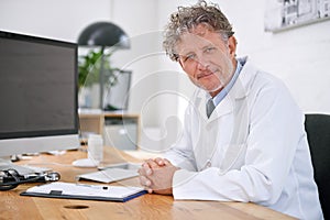 Tell me where it hurts. Portrait of a smiling mature doctor sitting at a desk in his office.