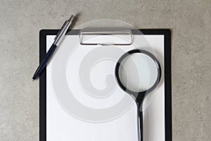 Template of white paper with a ballpoint pen and magnifying glass on light grey concrete background in a black tablet with a clip