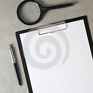 Template of white paper with a ballpoint pen and magnifying glass on light grey concrete background in a black tablet with a clip