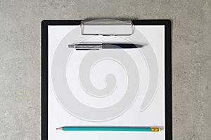 Template of white paper with a ballpoint pen and simple pencil on light grey concrete background in a black tablet with a clip.