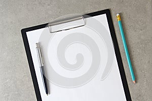 Template of white paper with a ballpoint pen and simple pencil on light grey concrete background in a black tablet with a clip.